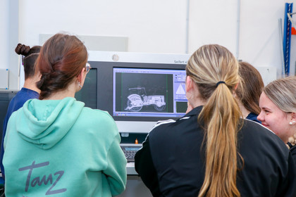 Rückansicht von vier Mädchen und einem Mann vor einem Monitor, auf dem ein Röntgenbild eines technischen Bauteils zu sehen ist.