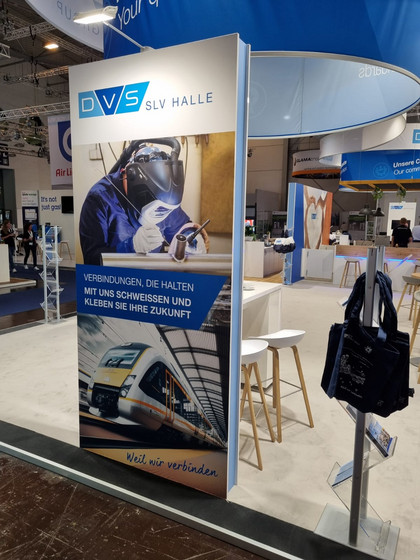 Messestand der DVS SLV Halle mit Roll-up-Banner, darauf ein Schweißer bei der Arbeit und ein Zugmotiv, dazu der Slogan: „Verbindungen, die halten – mit uns schweißen und kleben Sie Ihre Zukunft. Weil wir verbinden“.