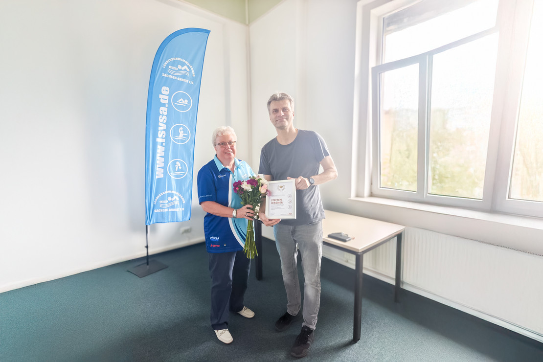 Eine ältere Frau im blauen Vereinsshirt überreicht einem Mann in Freizeitkleidung einen Blumenstrauß und eine Urkunde; im Hintergrund steht ein blauer Roll-Up des Landesschwimmverbandes Sachsen-Anhalt e. V.