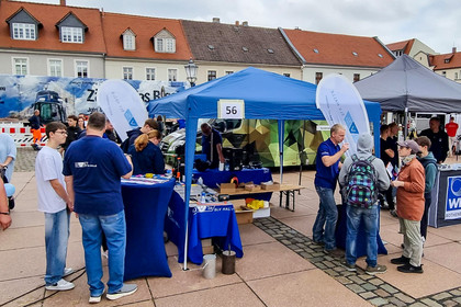 Zahlreiche Personen besuchen Informationsstände auf einem Platz im Freien. Im Mittelpunkt steht ein blauer SLV Halle Pavillon mit der Nummer 56, an dessen Tischen Besucher Informationsmaterial und Ausstellungsstücke betrachten.