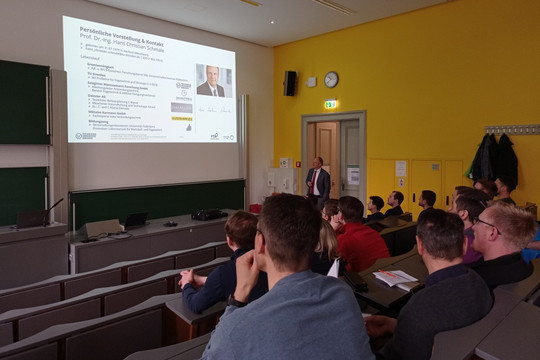 Ein Mann steht in einem Hörsaal vor einer Projektion mit dem Titel „Studienbegleitende Qualifizierung zum SFI – Eine Erfolgsgeschichte“ und spricht zu einem Publikum aus etwa zehn Personen, das aufmerksam zuhört.