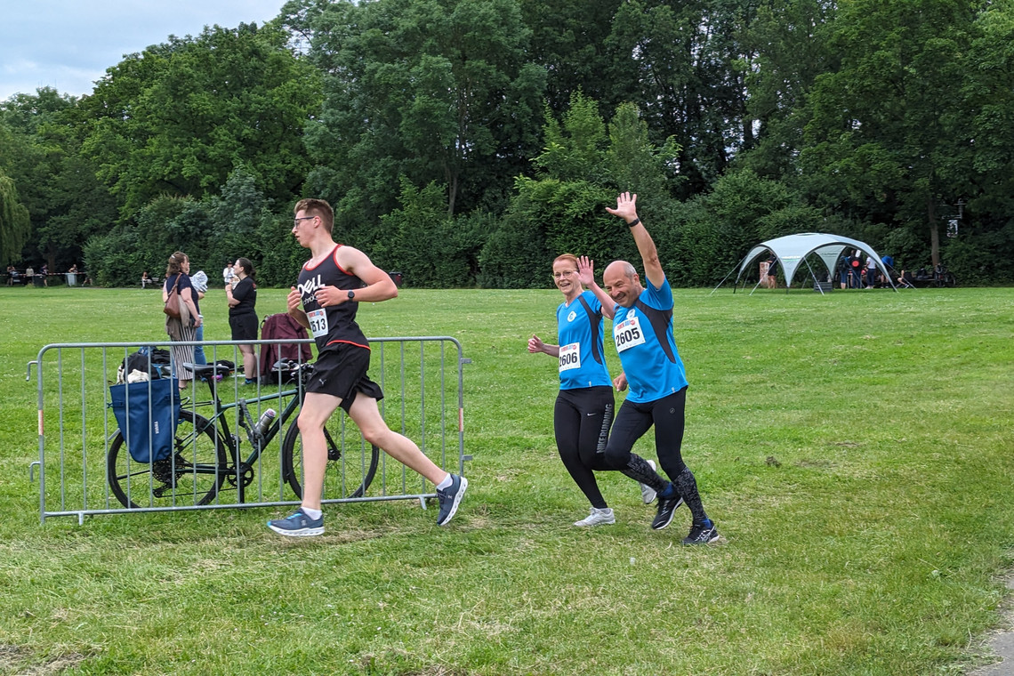 Zwei Personen in blauen Firmenlauf-Shirts laufen lächelnd und winkend nebeneinander über eine Wiese. Vor ihnen joggt ein Mann in schwarzem Laufshirt. Im Hintergrund stehen Zuschauende, Fahrräder und ein weißes Pavillonzelt.