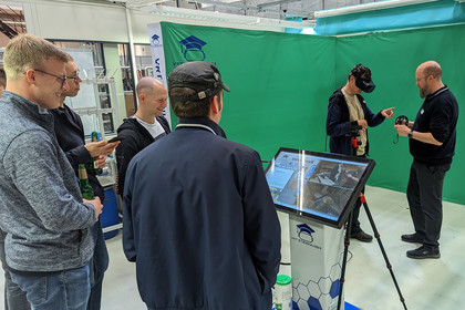 Mehrere Männer stehen in einer grünen VR-Trainingsumgebung, einer trägt ein Headset, ein anderer reicht ihm ein Controller, Bildschirm zeigt Schweißsimulation
