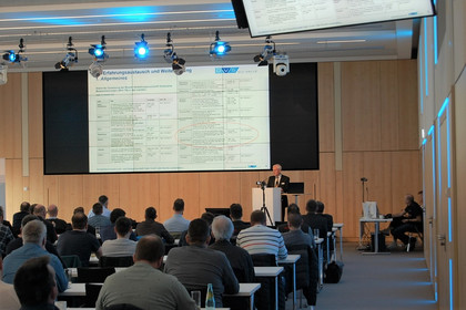 Teilnehmende sitzen in einem modernen Konferenzraum und verfolgen eine Präsentation mit Tabelle auf großer Leinwand, Redner steht am Pult, blaue Deckenbeleuchtung