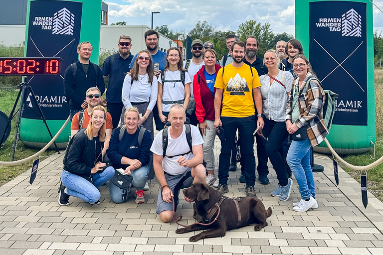 Eine größere Gruppe von Personen steht und kniet gemeinsam im Freien auf einem sandigen Untergrund. Mehrere Personen tragen farbige Sportkleidung, links und rechts stehen dunkle Werbebanner mit weißen und grünen Grafikelementen.