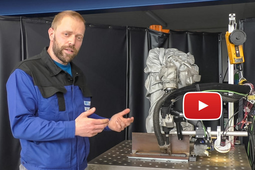 Ein Mann in blauer Arbeitskleidung steht vor einem Schweißroboter mit angeschlossener Technik und erklärt dessen Funktion, im Vordergrund ist ein eingeblendetes YouTube-Play-Symbol zu sehen.