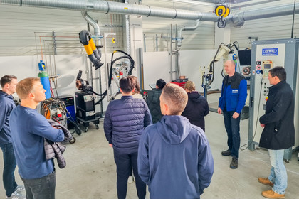 Sieben Personen stehen in einer Werkhalle und hören einem Mann zu, der technische Geräte und Schweißroboter erklärt.
