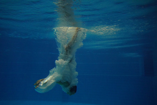 Eine Person taucht kopfüber ins Wasser und ist vollständig von einer weißen Blasenspur umgeben; das Bild ist unter Wasser aufgenommen und zeigt die blaue Kachelwand des Schwimmbeckens.