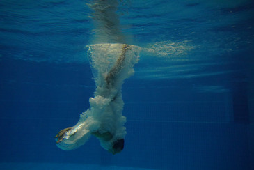 Eine Person taucht kopfüber ins Wasser und ist vollständig von einer weißen Blasenspur umgeben; das Bild ist unter Wasser aufgenommen und zeigt die blaue Kachelwand des Schwimmbeckens.