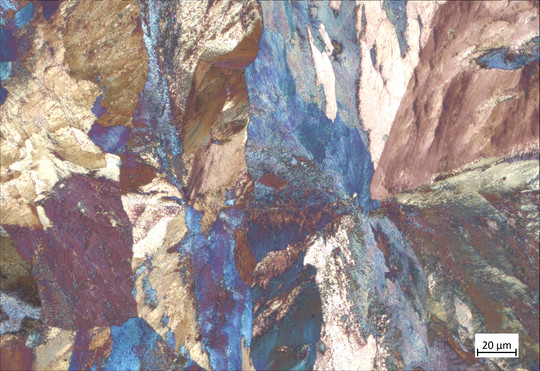 Mikroskopaufnahme eines metallischen Gefüges mit großflächigen kristallartigen Bereichen in Blau, Braun, Gold und Violett. Die Aufnahme zeigt unterschiedliche Gefügestrukturen mit feinen Linienmustern und enthält unten rechts einen Maßstab von 20 µm.