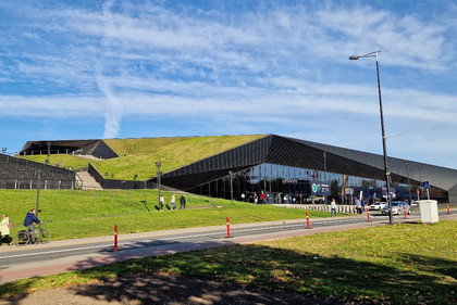 Moderne Messehalle mit begrünter Dachfläche und schwarzer Glasfassade, Menschen bewegen sich davor entlang eines Gehwegs bei sonnigem Wetter, blauer Himmel mit Schleierwolken.