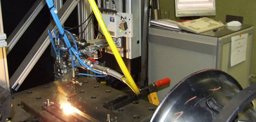 Eine automatisierte Schweißanlage mit Lasereinheit bearbeitet ein metallisches Werkstück auf einem Positioniertisch. Die Anlage ist mit blauen und gelben Leitungen verbunden, im Hintergrund steht ein Steuerungs-PC mit geöffnetem Messprogramm.