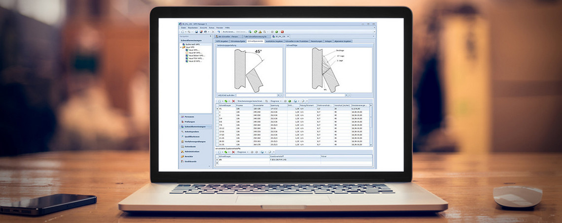 Ein geöffneter Laptop steht auf einem Holztisch, auf dem Bildschirm ist eine technische Software mit Schweißnahtdarstellungen und tabellarischen Daten zu sehen. Daneben liegen eine Computermaus und ein Smartphone, im Hintergrund ist eine Werkhalle unscharf erkennbar.