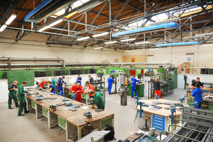 Mehrere Personen arbeiten in blauer, grüner und roter Arbeitskleidung an Werkbänken und Maschinen in einer großen, gut beleuchteten Werkhalle.