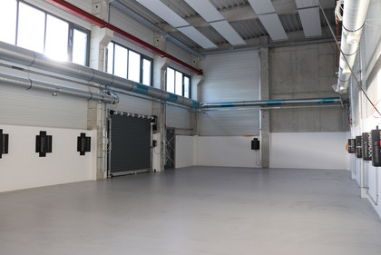 Leere, neu gebaute Industriehalle mit grauem Boden, Rolltor, Belüftungssystem und schwarzen Wandanschlüssen, hohe Fenster lassen Tageslicht herein