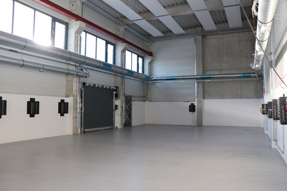 Leere, neu gebaute Industriehalle mit grauem Boden, Rolltor, Belüftungssystem und schwarzen Wandanschlüssen, hohe Fenster lassen Tageslicht herein