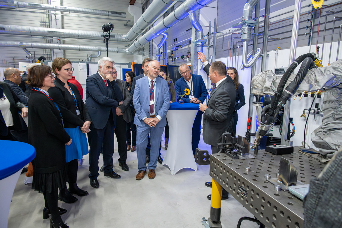 Eine Gruppe aus sieben Männern und fünf Frauen in Businesskleidung steht in einer Werkhalle um einen Vortragenden, der ein technisches Bauteil an einem Roboterschweißplatz erklärt; im Hintergrund sind Lüftungsanlagen und weitere Messebesucher:innen zu sehen.