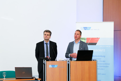Zwei Männer stehen nebeneinander an Rednerpulten mit SLV-Halle-Logo, einer spricht, der andere hört zu, rechts ein Roll-Up mit Informationen zur Weiterbildung im Korrosionsschutz