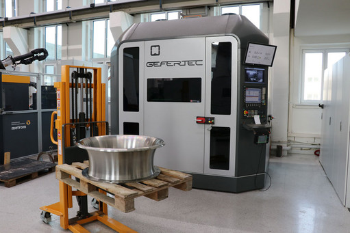 In einer modernen Werkhalle steht eine GEFERTEC-3D-Druckmaschine, davor ein Gabelhubwagen mit einer metallischen Einlauftrompete auf einer Holzpalette.