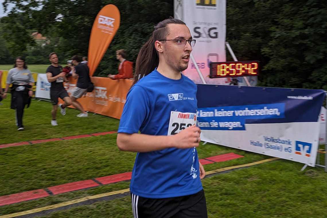 Ein Mann im blauen Trikot läuft mit Startnummer durchs Ziel eines Firmenlaufs in Halle. Im Hintergrund sind weitere Läufer\:innen, ein Zeitmessgerät und Werbebanner von Sponsoren zu sehen. Die Szene spielt sich auf einer grünen Wiese ab.