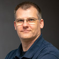 Matthias Schmidt, im dunkelblauen Hemd und Brille, lächelnd vor neutralem Hintergrund