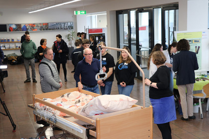  Jugendliche in schwarzen T-Shirts mit dem Logo „GWG“ präsentieren in elektrisch modifiziertes Pflegebett. Weitere Besuchende und Informationsstände sind im Hintergrund zu sehen.