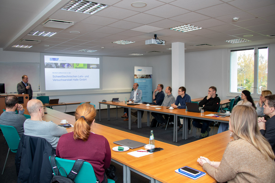 Ein Mann hält eine Präsentation vor einer sitzenden Gruppe aus sieben Frauen und acht Männern in einem hellen Konferenzraum; auf der Leinwand ist das Logo der Schweißtechnischen Lehr- und Versuchsanstalt Halle GmbH zu sehen.