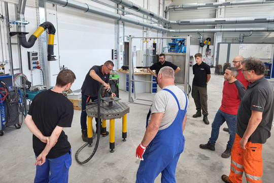 Eine Gruppe von Mitarbeitern beobachtet eine Vorführung zum induktiven Richten auf einem Metalltisch