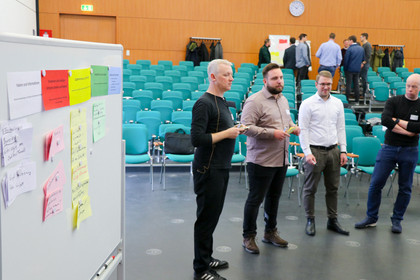 Vier Männer stehen in einem Workshopraum mit leeren Stuhlreihen vor einer Pinnwand mit farbigen Moderationskarten. Weitere Teilnehmer diskutieren im Hintergrund.