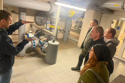 Ein Mann erklärt einer Gruppe aus vier Männern und einer Frau technische Rohrleitungen in einem Versorgungsraum; alle blicken aufmerksam auf das dargestellte System aus Ventilen, Armaturen und Absperrklappen.