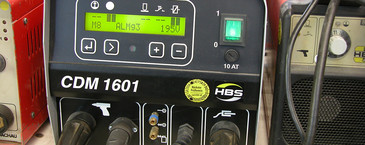 Ein CDM 1601 Bolzenschweißgerät von HBS mit grün leuchtendem Display und mehreren angeschlossenen Kabeln steht auf einer Werkbank. Das Display zeigt Einstellungen wie „M8 ALMg3“ und „19.5 V“. Oben liegt ein Schweißpistolenaufsatz.