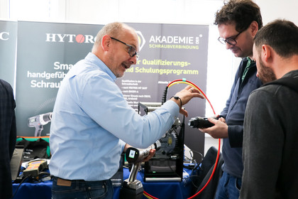 Ein Aussteller zeigt zwei Männern ein technisches Gerät mit rotem Kabel, im Hintergrund Banner der Akademie der Schraubverbindung.
