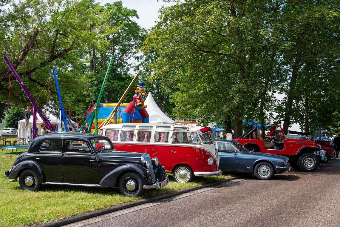 Mehrere Oldtimer und klassische Fahrzeuge stehen auf einer Wiese unter Bäumen. Im Hintergrund sind ein weißes Festzelt, weitere Besucher und Veranstaltungsstände im Freien zu sehen.