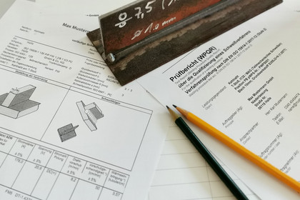 uf einem Tisch liegen Prüfberichte mit technischen Zeichnungen und Tabellen, daneben ein gelber und ein schwarzer Bleistift sowie ein beschriftetes Schweißprobestück mit Nahtbewertung.