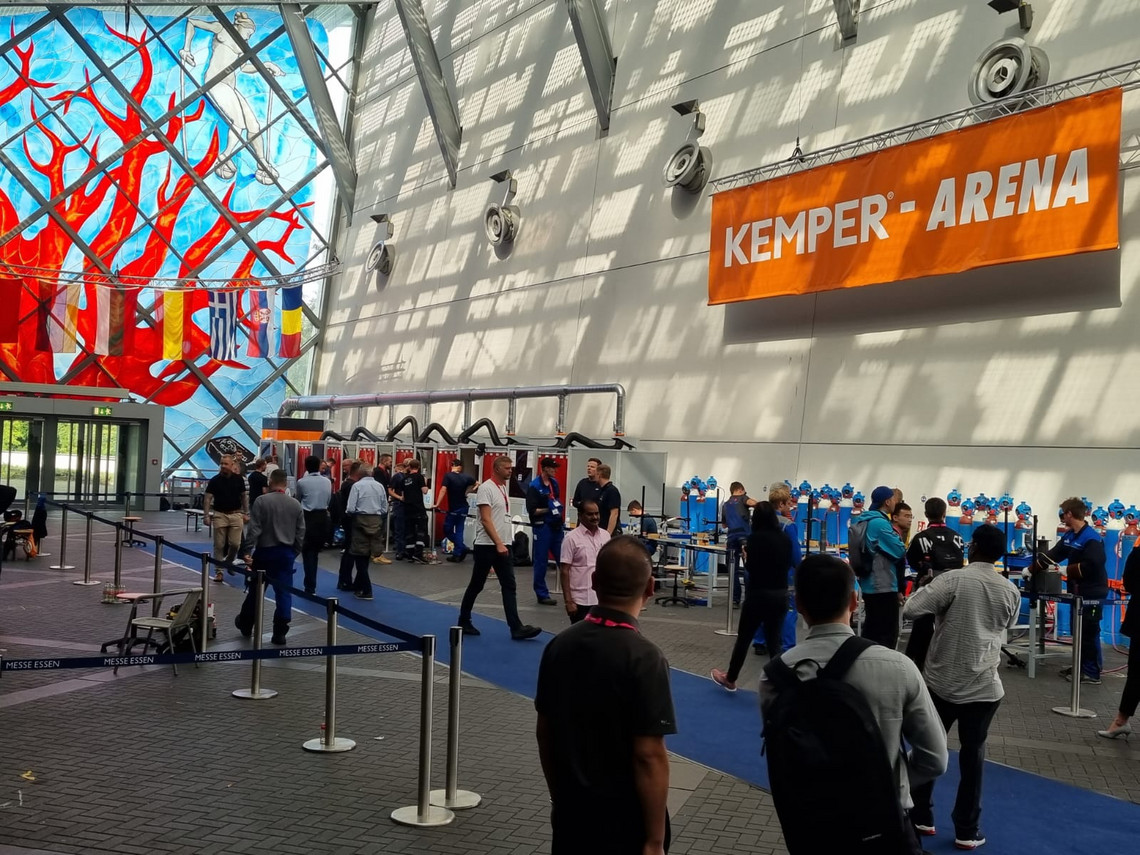 Menschen betreten eine große Messehalle mit dem Schild „KEMPER ARENA“, im Hintergrund ist ein großes Wandbild mit Baummotiv und Nationalfahnen zu sehen; die Halle ist durch Abtrennungen strukturiert und mit blauen Spinden und Lüftungssystemen ausgestattet.