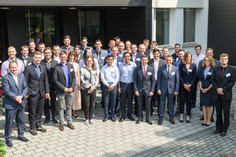 Gruppenfoto von 40 Personen in Businesskleidung vor einem Gebäude; darunter 33 Männer und 7 Frauen. Alle blicken in die Kamera und stehen auf einem gepflasterten Vorplatz.