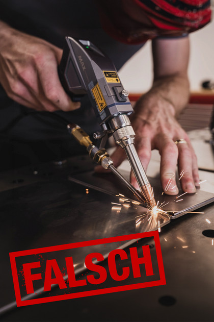Eine Person führt Schweißarbeiten an einem Metallbauteil auf einer Werkbank aus. Im Vordergrund ist der rote Schriftzug „FALSCH“ zu sehen, die Person trägt keine erkennbaren Schutzhandschuhe.