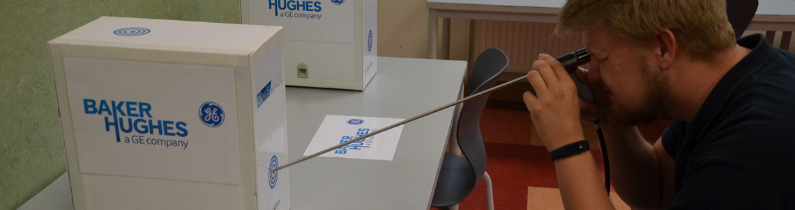 Ein junger Mann betrachtet mit einem Endoskop ein Prüfobjekt in einem weißen Gehäuse mit dem Logo von Baker Hughes; er kniet an einem Tisch in einem Schulungsraum.