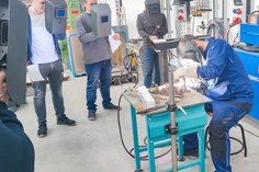 Ein Ausbilder demonstriert eine Schweißtechnik an einem Werkstück auf einem Tisch, während fünf Teilnehmende mit Schweißschilden und Unterlagen im Halbkreis zuschauen. Im Hintergrund stehen technische Geräte und Gasflaschen.