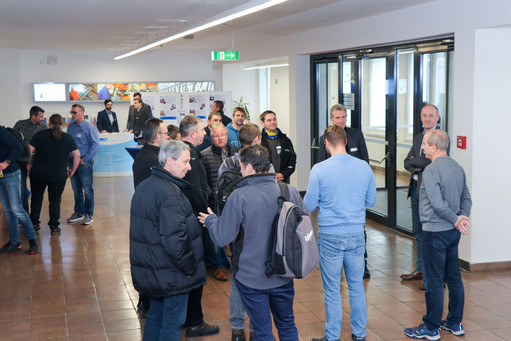 Mehrere Männer stehen in kleinen Gruppen im Eingangsbereich eines modernen Veranstaltungsgebäudes und unterhalten sich; im Hintergrund ist ein Infotresen mit Roll-ups und Personal zu sehen.