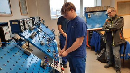 Eine auszubildende Person in blauer Arbeitskleidung arbeitet an einem pneumatischen Trainingssystem mit schräg gestellter Lochplatte; zwei weitere Personen beobachten die Durchführung im Schulungsraum.