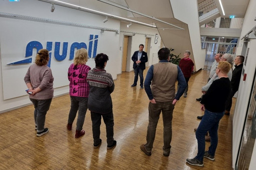 Elf Personen stehen in einem Flur mit Holzfußboden und hören einem Mann in Anzug zu, der vor dem großen Wandlogo „njumii“ spricht; die Gruppe befindet sich in einem modernen Bildungs- oder Verwaltungsgebäude.