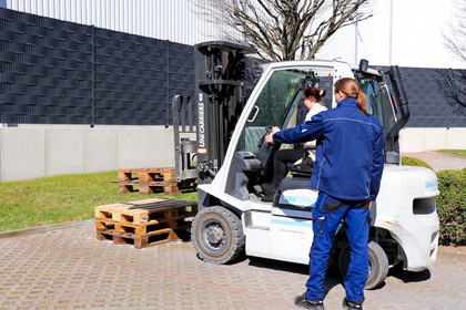 Eine junge Frau sitzt in einem weißen Gabelstapler und übt das Anheben von Paletten, daneben steht ein Mann in blauer Arbeitskleidung und gibt Anweisungen.