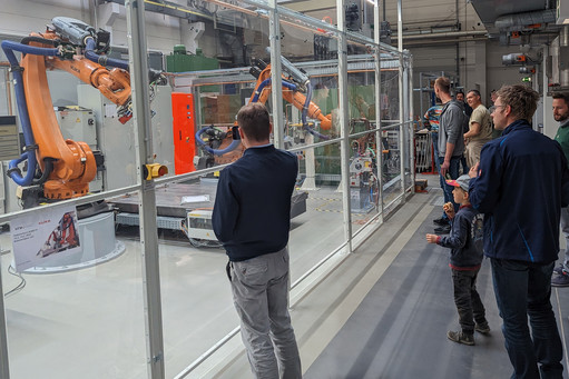 Männer und ein Kind betrachten durch Glasscheibe zwei orangefarbene KUKA-Roboter beim Schweißen, dahinter moderne Technik und Hallenstruktur sichtbar