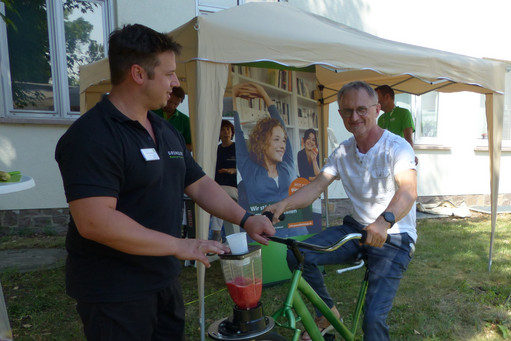 Ein Mann in Jeans und weißem T-Shirt tritt auf einem grünen Smoothie-Fahrrad in die Pedale, während ein Betreuer in schwarzer Kleidung das Gerät festhält. Im Hintergrund stehen weitere Personen unter einem Pavillon.