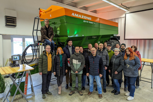 Gruppe von 20 Personen, 14 Männer und 6 Frauen, steht vor einem großen grün-orangefarbenen AMAZONE-Streuer in einer Halle, eine Person sitzt oben auf dem Fahrzeug
