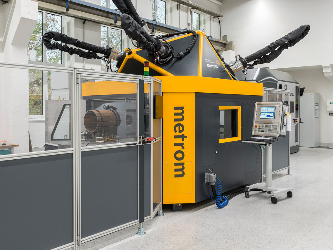 Gelb-schwarze Hybridmaschine von metrom mit mehreren Roboterarmen steht in einer abgetrennten Werkzelle; durch ein Fenster ist ein zylindrisches Werkstück erkennbar, rechts befindet sich das Bedienpult mit Monitor.