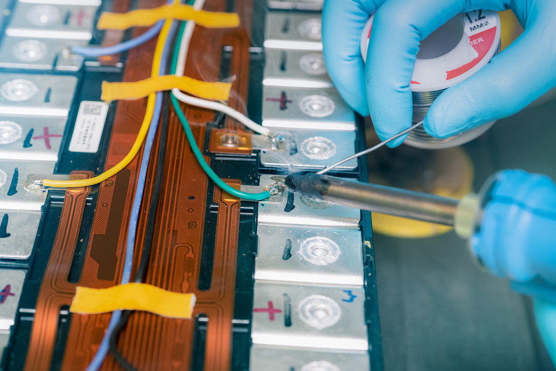 Nahaufnahme einer Person mit blauen Schutzhandschuhen beim Löten an einem Batteriepack, mehrere Zellen, Kabel und flexible Leiterbahnen sind sichtbar.