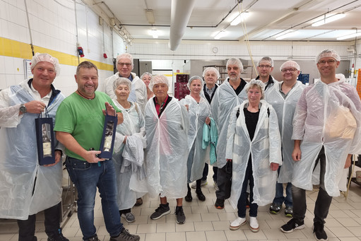 Vier Frauen und zehn Männer in Hygiene-Schutzkleidung stehen in einer Großküche oder lebensmittelverarbeitenden Betriebsstätte, zwei von ihnen halten Weinpräsente, alle blicken lächelnd in die Kamera.