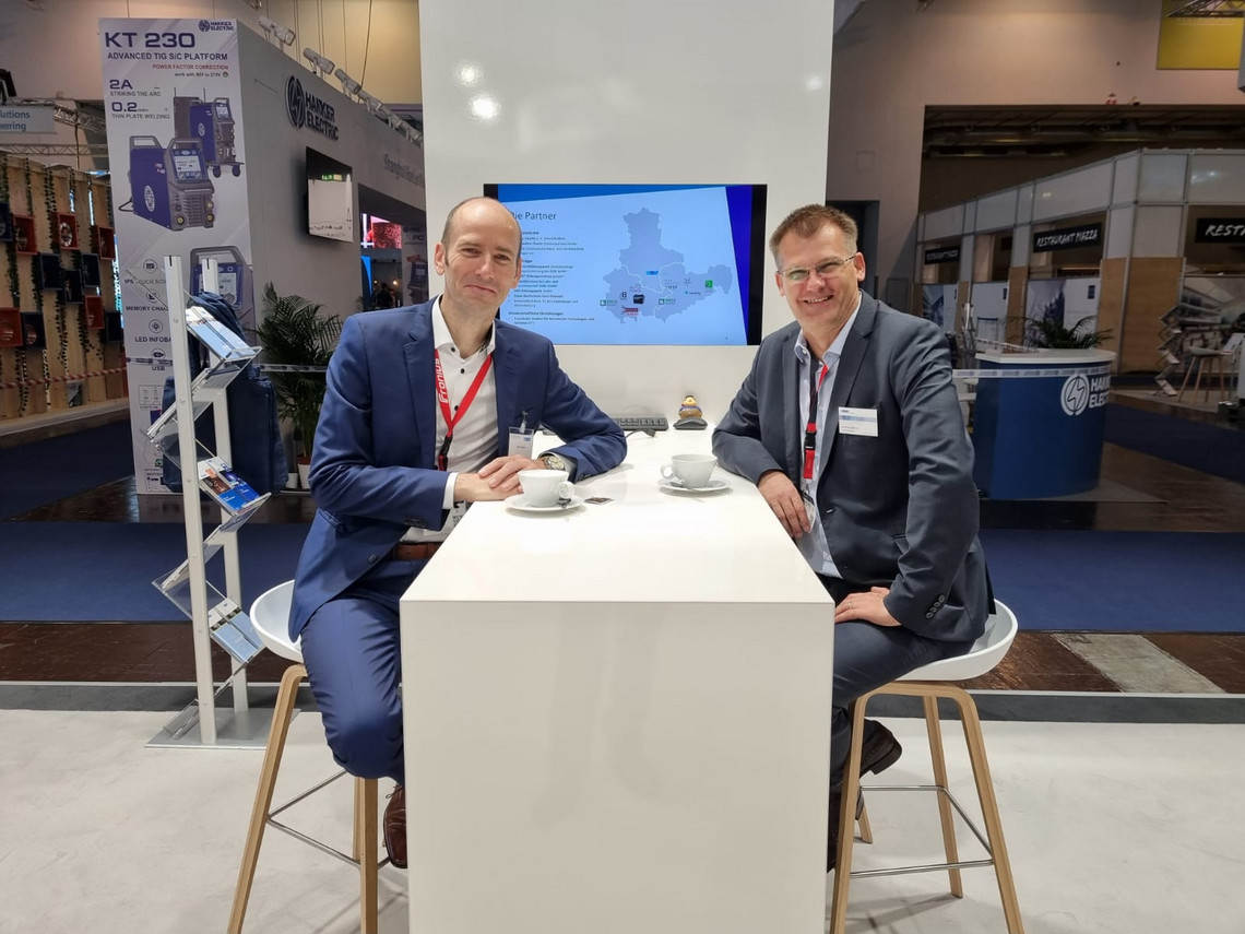 Zwei Männer in Anzügen sitzen an einem weißen Stehtisch mit Kaffeetassen auf einer Messe, im Hintergrund ist ein Monitor mit einer Karte sowie ein Stand mit Schweißtechnik zu sehen.